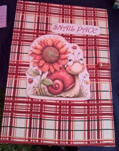 Decorative card showing a sunflower and a smiling snail on a red plaid background, with the text'SHAIL PACE' at the top.