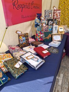 740128661_27052389611124018_1371663851847647412_n | Busy Bees Patchwork Vendor table of handmade notebooks with colorful covers and price tags at a market stall, pink banner in back reading'Rosepetals Creations'.