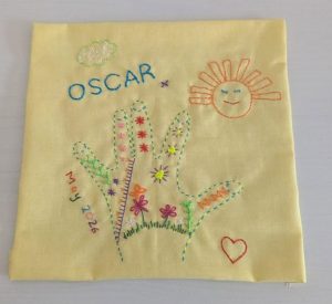 Embroidered yellow cloth with two colorful hands, flowers, sun, cloud, heart, and the name 'OSCAR'—a child's art piece.
