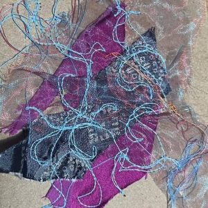 Tangled blue thread draped over purple and black lace fabric scraps on a beige carpet.