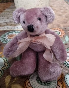 729293596_26930092146687099_4579059709652052817_n | Busy Bees Patchwork Lavender plush teddy bear with a pink ribbon bow resting on patterned fabric.