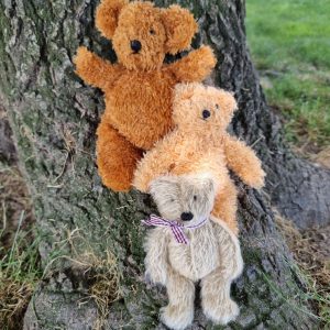 Three fluffy teddy bears stacked on a tree trunk outdoors, one wearing a striped scarf in the foreground while grass and trees are in view behind them.