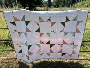 739276548_27050588154637497_8954961844036323989_n | Busy Bees Patchwork White patchwork quilt with green, rust, and floral triangle blocks hangs over a metal fence in a sunlit field.