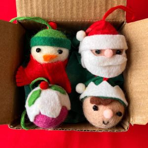 Festive felt Christmas ornaments in a cardboard box: a snowman with green hat and scarf beside a Santa head with white beard and red hat, on a red surface at the bottom.