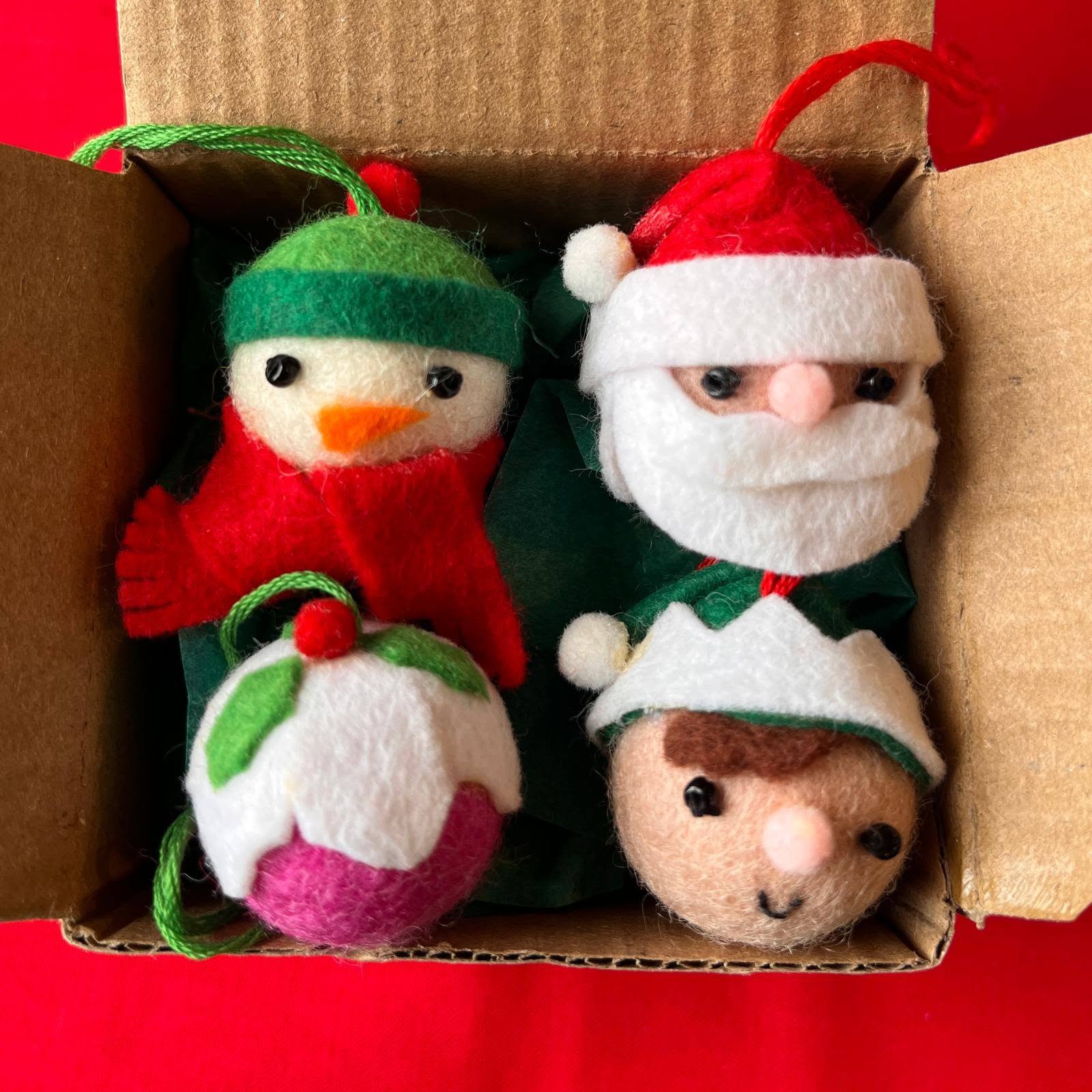 749269412_27134687482894230_7997739706519892106_n | Busy Bees Patchwork Festive felt Christmas ornaments in a cardboard box: a snowman with green hat and scarf beside a Santa head with white beard and red hat, on a red surface at the bottom.