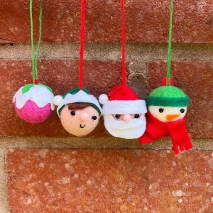 749542542_27134687122894266_4551868300570885620_n | Busy Bees Patchwork Felt Christmas ornaments: a pink-and-green ball, a Santa head, a snowman, and an elf hanging by red and green cords against a brick wall.