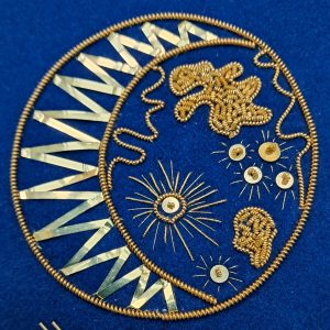 Close-up of gold bead embroidery on royal blue fabric, showing circular medallions with radiating spokes and a scalloped border with metallic triangles on the left.