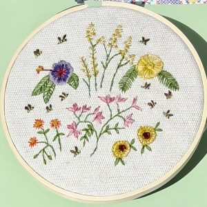 Circular mosaic floral embroidery on a pale green wall, featuring yellow, pink, purple flowers with green stems inside a beige-framed round artwork.