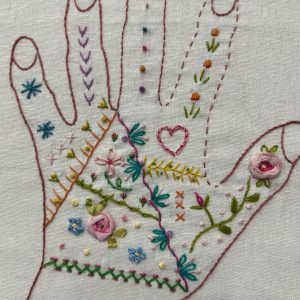 Embroidery of a hand outline with colorful stitches and floral motifs on light fabric, with a wooden frame edge visible at bottom.