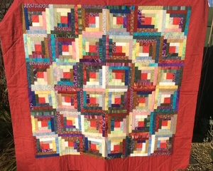 Colorful patchwork quilt with a red border hanging outdoors, showing interlocking square blocks in many colors.