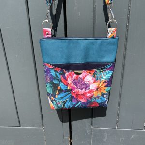 Teal handbag with a vibrant floral front hangs on a gray wooden door, showing metal clasps on colorful straps.