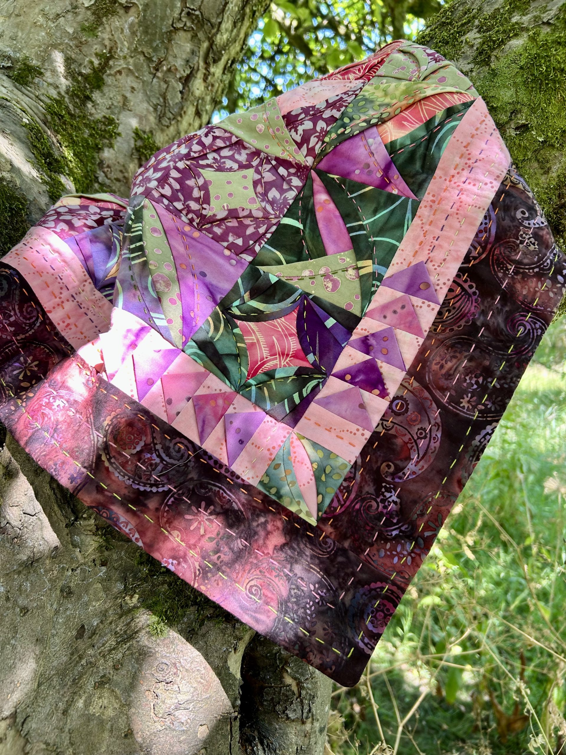 IMG_1715 | Busy Bees Patchwork Colorful patchwork quilt draped over a mossy tree trunk in a sunlit outdoor setting.