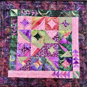 Colorful patchwork quilt with purple, pink, green squares and decorative stitching, hanging outdoors on a wooden surface.