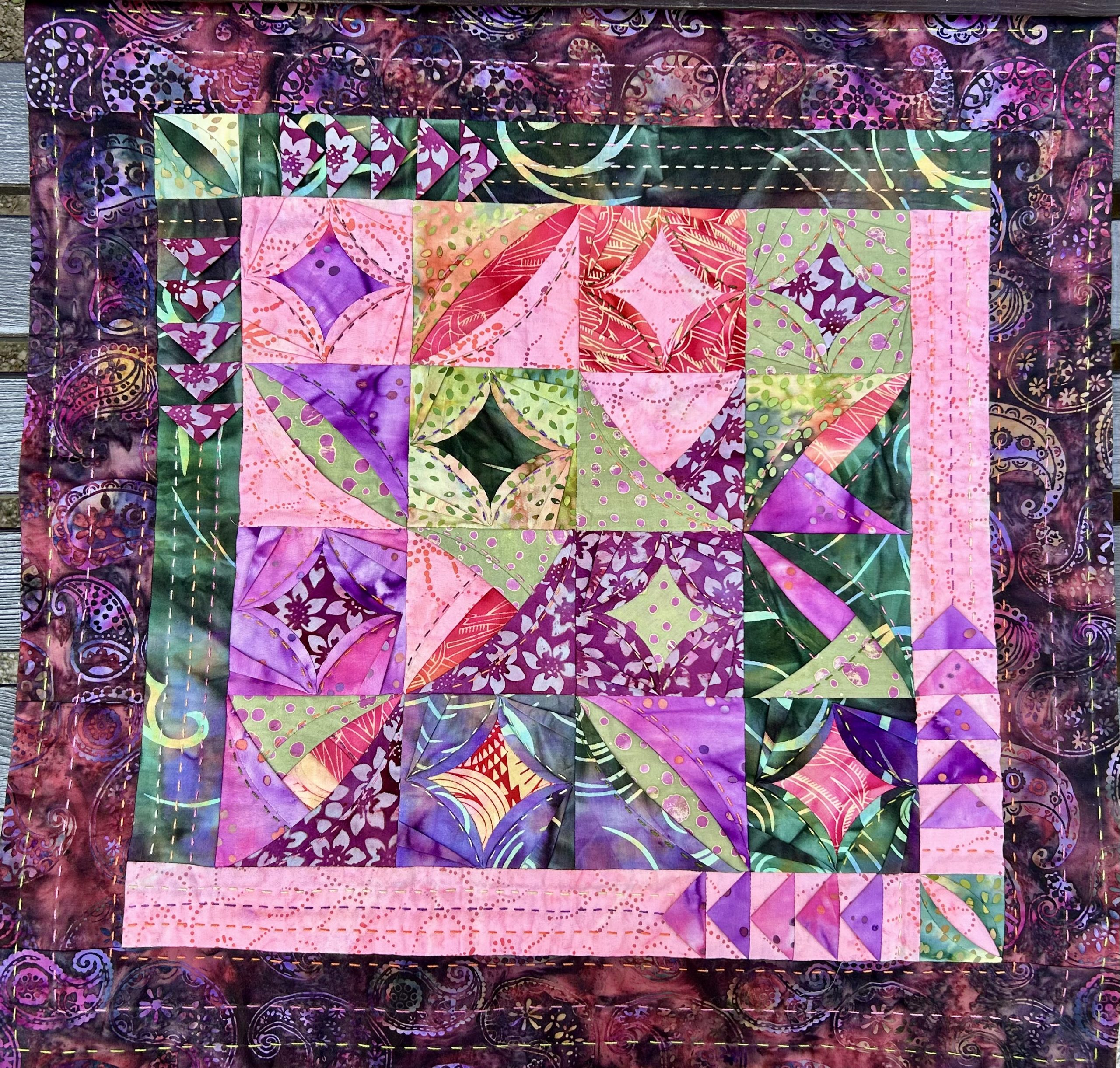 IMG_1719 | Busy Bees Patchwork Colorful patchwork quilt with purple, pink, green squares and decorative stitching, hanging outdoors on a wooden surface.