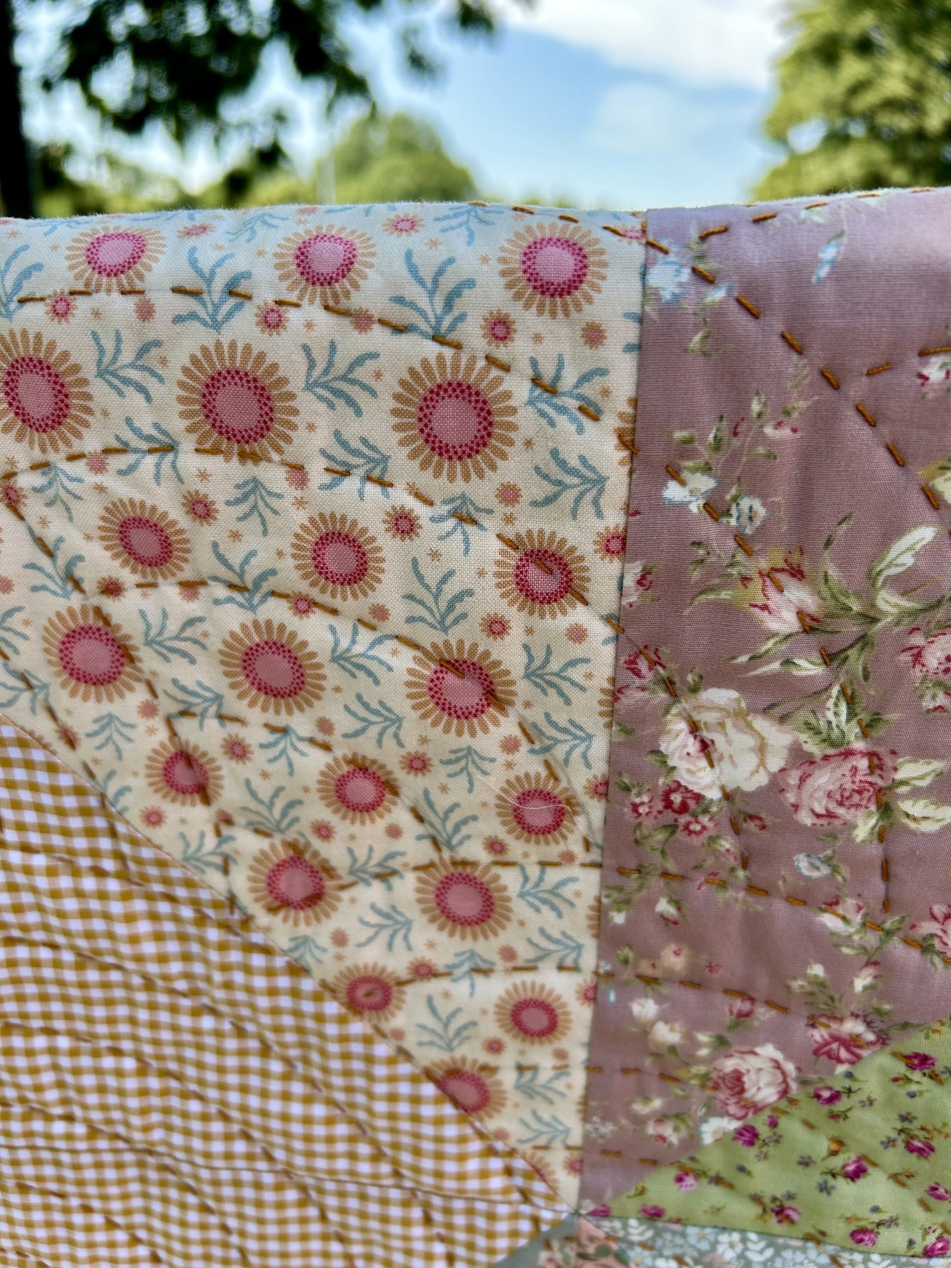 IMG_1723 | Busy Bees Patchwork Close-up of a colorful patchwork quilt with floral, gingham, and pastel fabrics stitched together, stitching visible, outdoor greenery and blue sky blurred in the background
