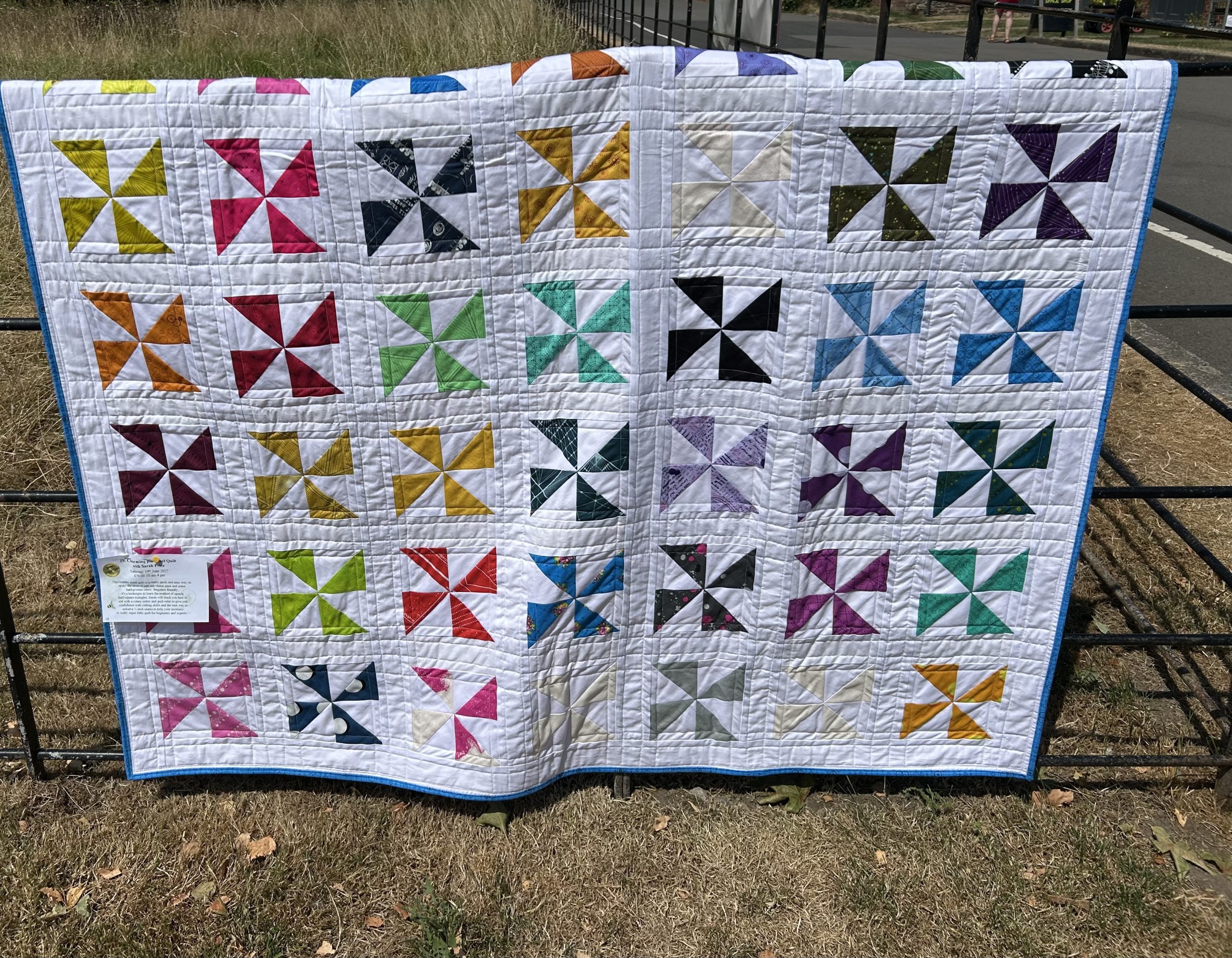IMG_1726 | Busy Bees Patchwork Patchwork quilt with colorful pinwheel blocks draped over a black metal fence outdoors, blue binding along edges.