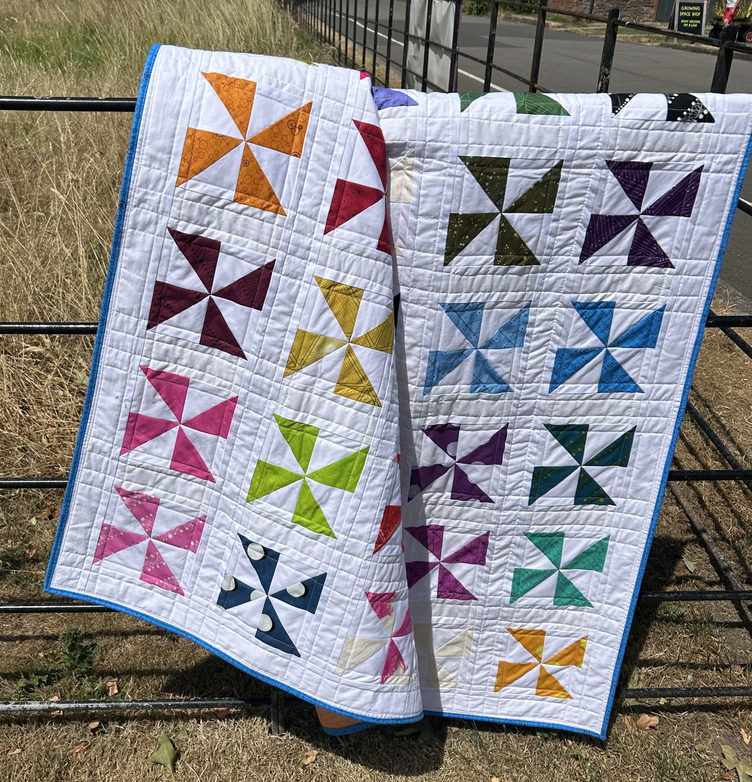 IMG_1727 | Busy Bees Patchwork Two colorful patchwork quilts with pinwheel blocks draped over a metal fence in a sunny, grassy area.