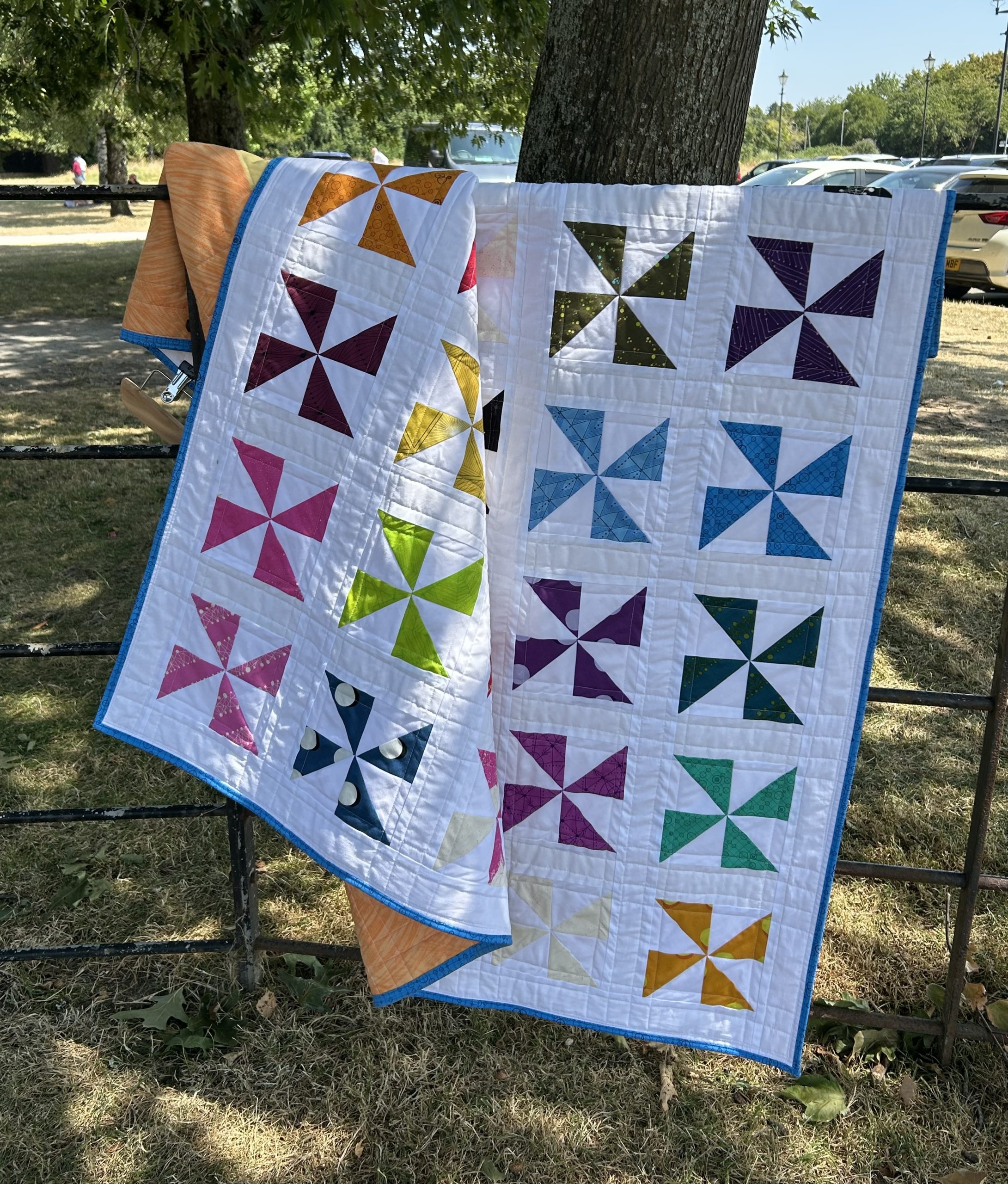 IMG_1729 | Busy Bees Patchwork Two patchwork quilts with colorful pinwheel blocks draped over a metal fence outdoors, sunlight filtering through trees in a park setting.