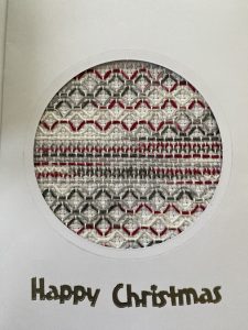 White card with a circular window revealing a red and gray stitched fabric pattern; visible text reads'Happy Christmas'.