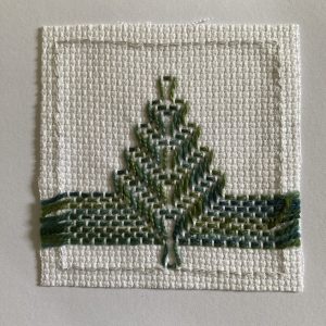 Small white fabric swatch with green cross-stitched tree above blue-green water/ridge.