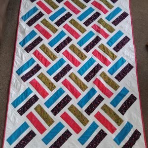 Colorful patchwork quilt spread on the floor with diagonal stripes in blue, pink, olive floral, and dark floral on a white background with a pink border edge.