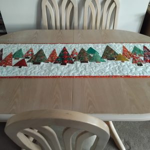 Quilted Christmas table runner with colorful fabric trees laid on a round wooden table, chairs in the background.