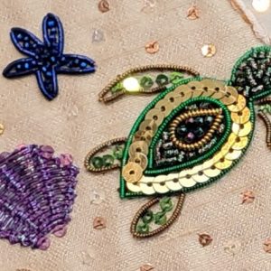 Beaded green and gold turtle appliqué on beige fabric with a blue starfish and purple shell nearby.